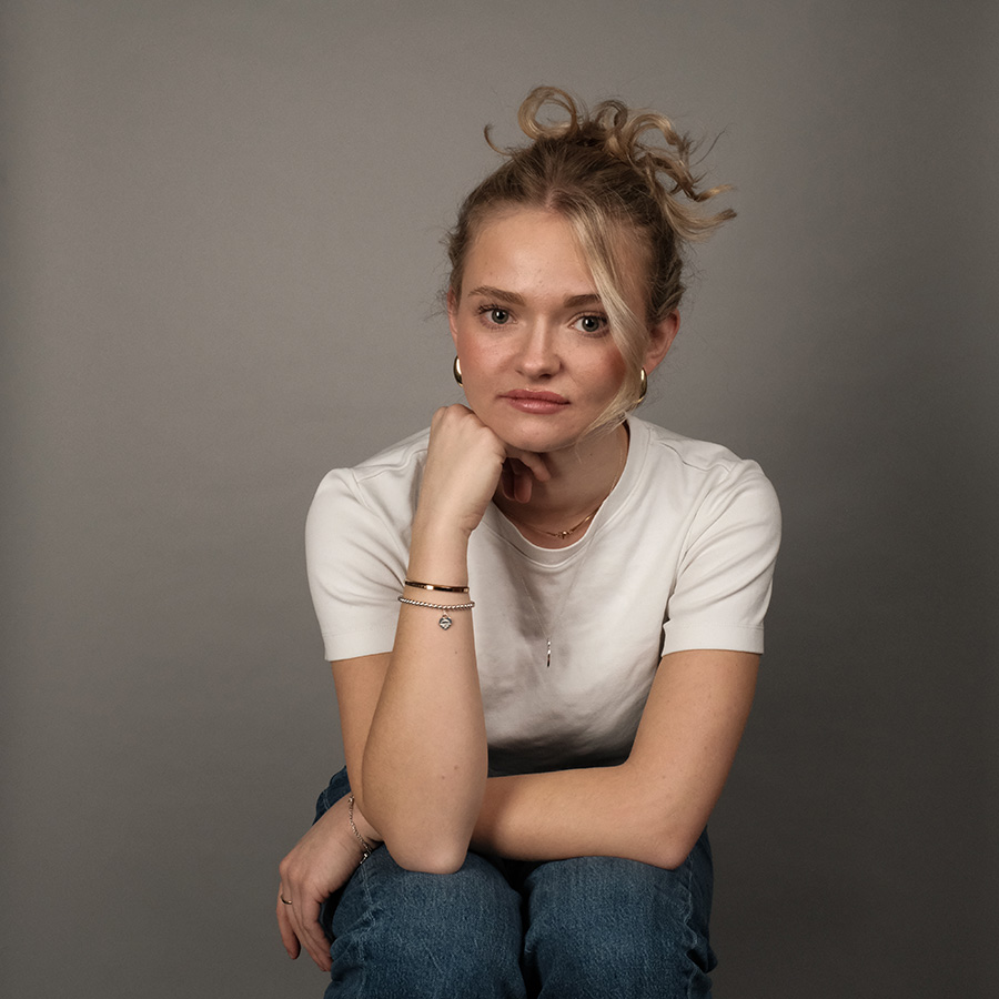 Portrait von Lina, Grafikdesign Studentin, sie trägt ein weißes tshirt und eine jeans, sie sitzt auf einem hocker und stützt ihren kopf in ihre hände, welche auf ihren knien stützen